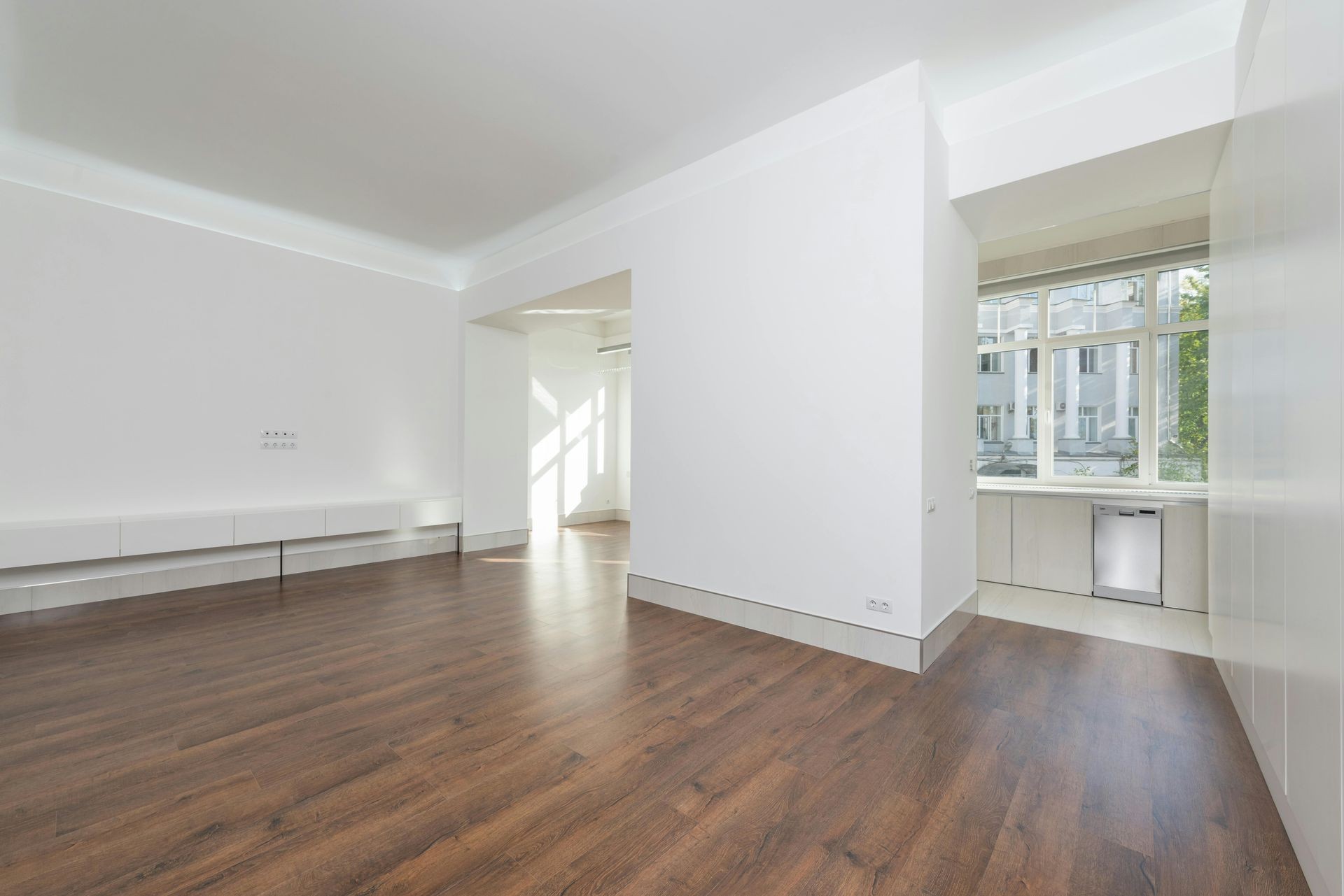 Spacious empty room with wooden flooring, white walls, and large windows offering natural light.