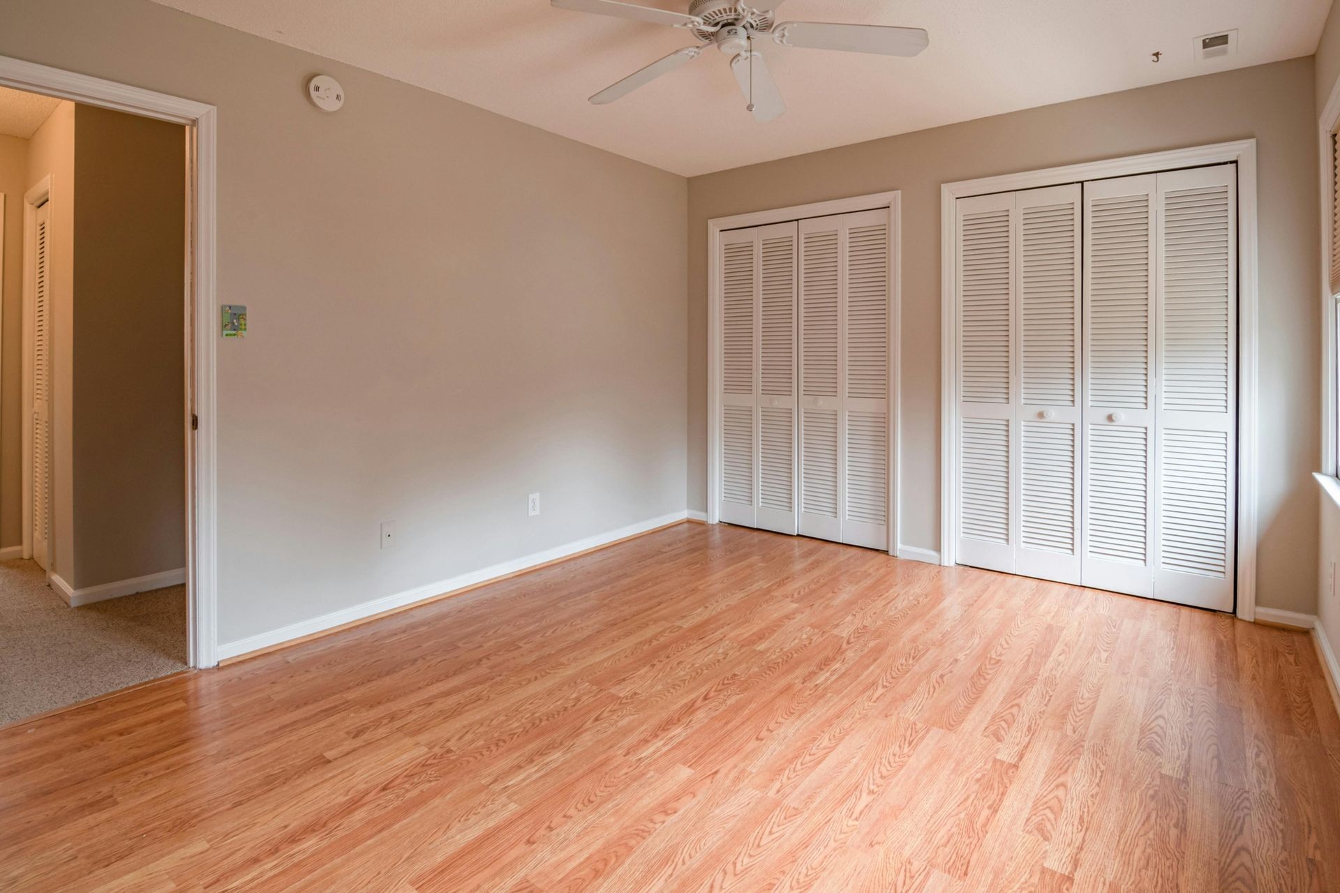 Empty room with light wood flooring, beige walls, ceiling fan, and two white closet doors.